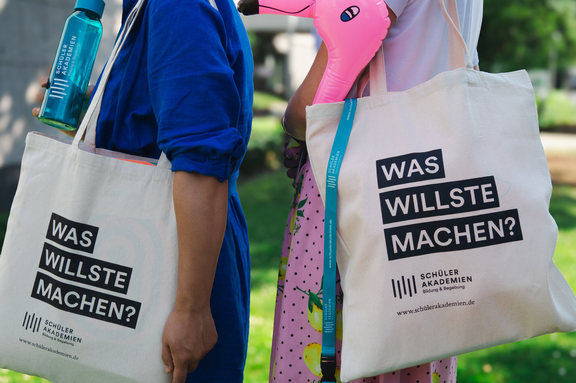Zwei Personen tragen eine Jutetasche mit dem Aufdruck "Was willste machen?" und dem Logo der Schülerakademie. Aus einer Tasche ragt ein rosa Aufblas-Flamingo. Die linke Person hält eine Trinkflasche mit dem Logo der Schülerakademie. 